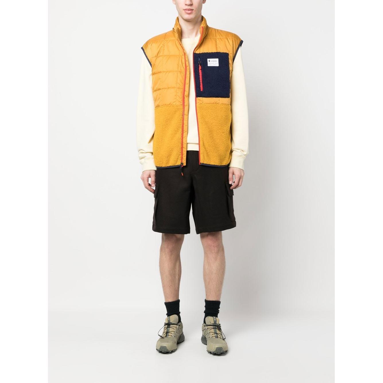 Model wearing Cotopaxi yellow padded gilet with chest logo patch, front zip, sleeveless design, over cream top and black shorts