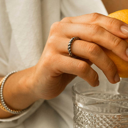 Close-up of a hand wearing AN JEWELS JEWELRY Mod. AAC.R05S-8 ring, holding an orange and resting on a glass.