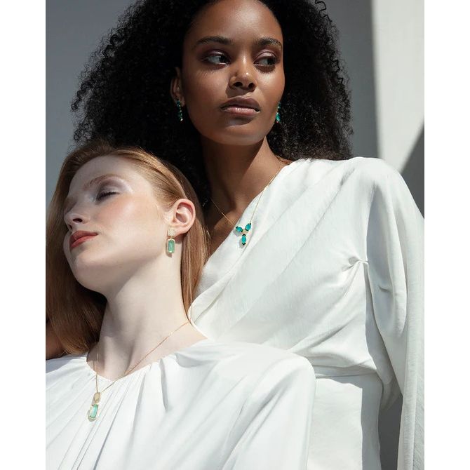 Two women wearing elegant white outfits and jewelry, showcasing necklaces and earrings with green gemstones in a stylish fashion setting.